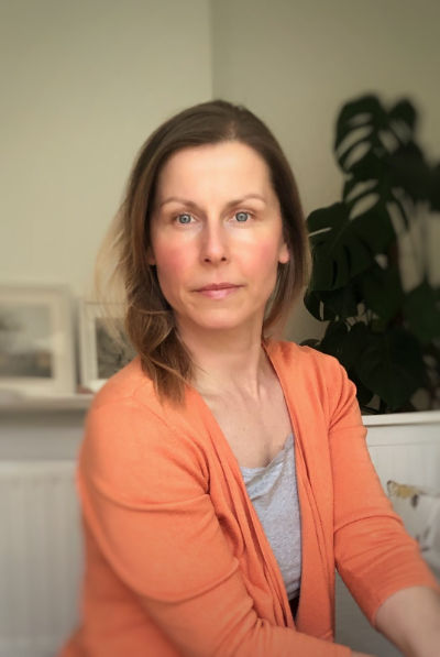 An image of Agi, a woman sitting on a chair with a plant in the background. She is calm
				and has a peaceful smile.